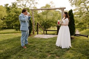 Emotional groom wipes tears seeing bride in white gown near rustic wooden wedding arch.