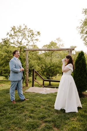 Bride photographs groom during outdoor first look at rustic wooden arch on lush green lawn.