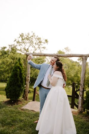 Bride and groom take a selfie together under a rustic wooden arch surrounded by lush greenery.