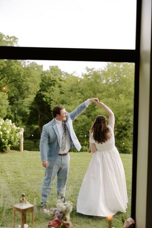 Groom spinning bride in white gown outdoors viewed through large window with garden backdrop.