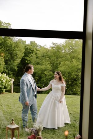 Bride and groom hold hands on green lawn viewed through large window at outdoor wedding venue.