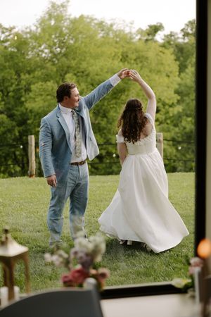 Groom twirls bride in flowing white dress on green lawn near glass venue doors.