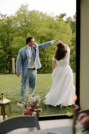 Groom in blue suit twirls bride in white gown near open glass doors with green landscape behind them.