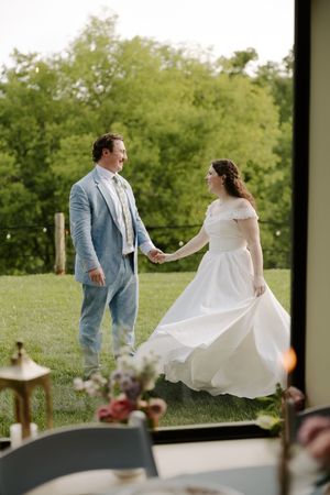 Newlyweds hold hands and smile at each other on green lawn, bride in flowing white dress and groom in light blue suit.