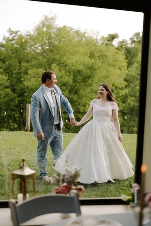Laughing bride and groom holding hands walking through green outdoor wedding venue.