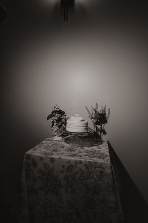 Elegant black and white photo of single-tier wedding cake with floral arrangements on patterned tablecloth.