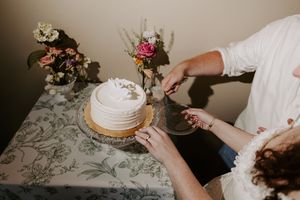 Bride and groom cutting elegant white wedding cake decorated with fresh flowers on floral tablecloth.