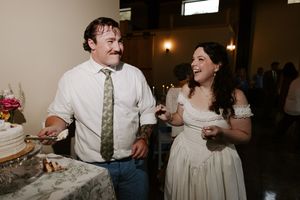 Bride in off-shoulder gown laughs with guest in white shirt near wedding cake at reception.