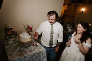 Groom cuts white wedding cake while bride watches smiling at elegant indoor reception.
