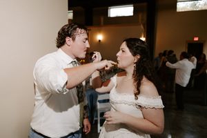 A groom in a white shirt feeds wedding cake to his bride in an off-shoulder dress at their reception.