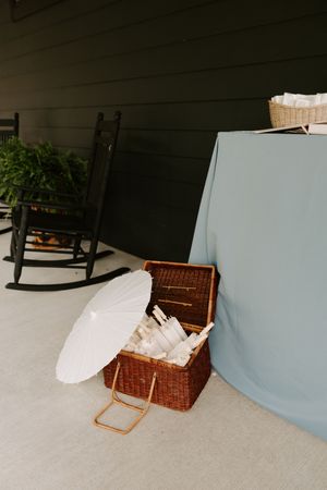 Wedding welcome table with wicker basket, white parasol, and programs displayed on a covered outdoor porch.