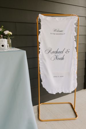 Welcome sign for Rachael and Noah wedding on June 27 2024 displayed on a gold stand with ruffled white fabric.