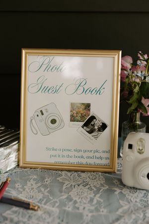 Gold-framed Photo Guest Book sign with Instax camera illustration at a wedding reception table with flowers.