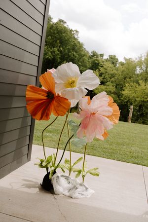 Large decorative paper flowers in orange, white, and pink displayed on a concrete step outside a barn venue.