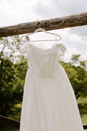 Elegant white off-shoulder lace wedding dress hanging on a rustic wooden beam against a cloudy sky.