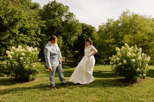 Bride in white gown and groom in blue suit walk among blooming hydrangeas in garden.