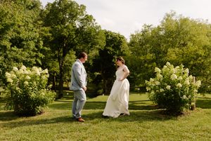 Bride and groom facing each other on a green lawn beside blooming white hydrangea bushes outdoors.