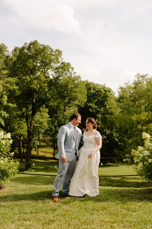Newlywed couple sharing a romantic moment while walking together on green grass surrounded by trees.