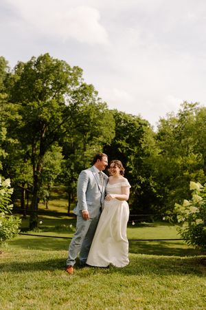 Bride and groom embracing and smiling outdoors on a sunny day with lush green trees behind them.