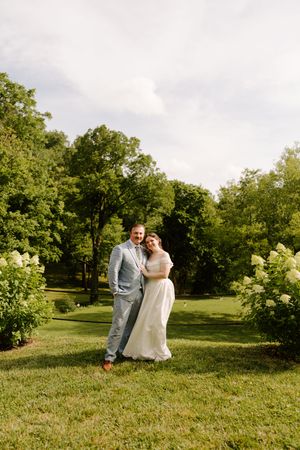 A bride and groom pose together on a lush green lawn surrounded by blooming white hydrangeas on their wedding day.
