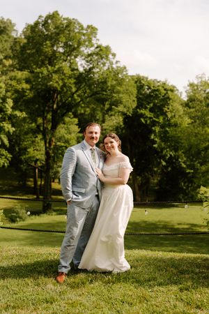 Bride and groom smile together in lush green outdoor setting, bride in off-shoulder gown, groom in blue suit.