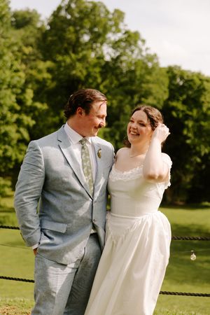 Smiling bride and groom pose together outdoors, with the groom in a light blue suit and bride in a lace dress.