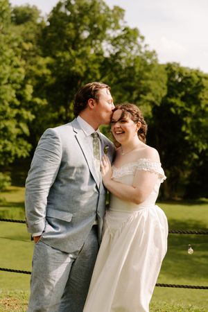 Groom in light blue suit kissing smiling bride on the cheek outdoors in a lush green garden setting.