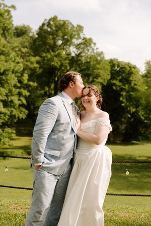 Groom kisses bride on cheek as she smiles in sunny outdoor garden setting with string lights.