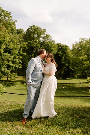 Bride and groom share a romantic kiss outdoors on lush green grass surrounded by trees on their wedding day.