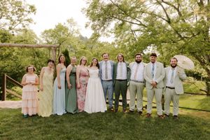 Wedding party poses together in colorful attire on a green lawn surrounded by lush trees.