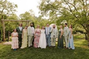 Wedding party poses outdoors on green lawn surrounded by trees, wearing elegant formal attire in pastel colors.