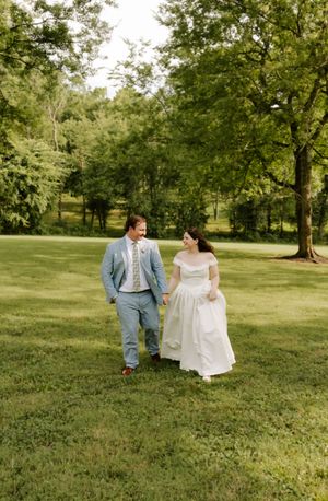 A laughing bride in an off-shoulder gown and groom in a light blue suit walk hand in hand across a green lawn.