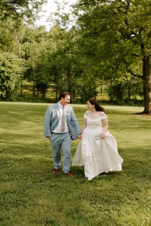 Smiling bride and groom hold hands walking through lush green park on summer wedding day.