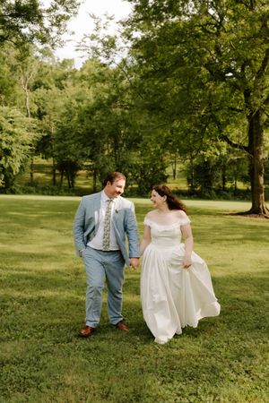 Happy newlywed couple walks hand in hand across green lawn surrounded by trees on wedding day.