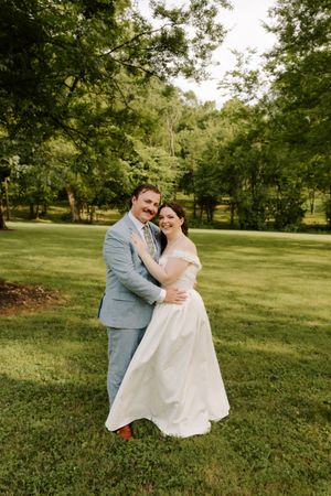 Groom embraces bride from behind as both smile in lush green garden, bride in off-shoulder wedding gown.