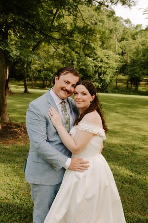Bride and groom embracing and smiling in a lush green outdoor setting, bride in off-shoulder white gown, groom in light blue suit.