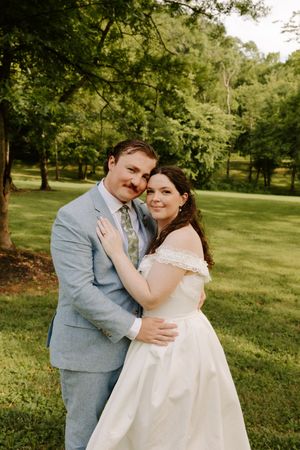 Bride and groom embrace outdoors on lush green grass surrounded by trees in summer wedding portraits.