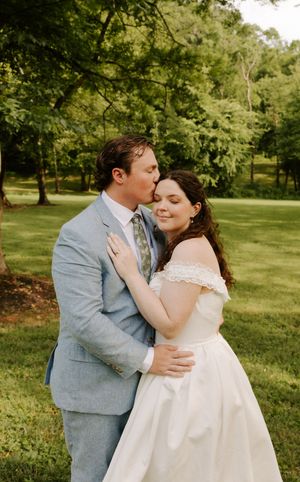 Groom in light blue suit kisses bride on cheek as she smiles in off-shoulder white wedding dress outdoors.