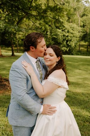 Groom in light blue suit tenderly kissing bride on cheek during outdoor wedding portrait session.