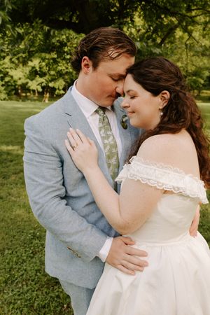 Newlyweds share an intimate forehead-to-forehead moment, bride in off-shoulder lace gown, groom in light blue suit.