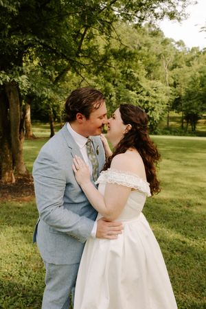 Bride and groom share a tender nose-to-nose moment while embracing beneath lush green trees outdoors.
