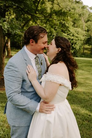 Bride and groom sharing a tender almost-kiss embrace outdoors surrounded by green trees on their wedding day.