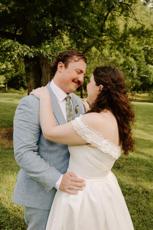 Smiling bride and groom embrace outdoors in lush green park, she in off-shoulder white gown, he in light blue suit.