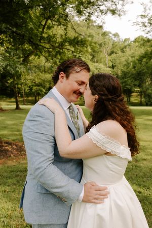 Bride and groom share a romantic forehead touch in a lush green park, groom in light blue suit, bride in off-shoulder gown.
