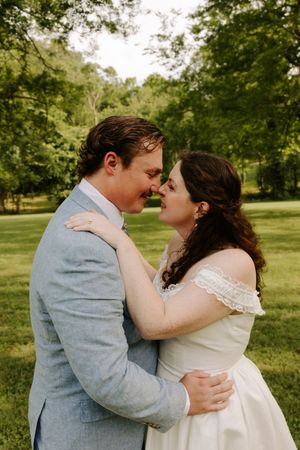 Newlyweds share an intimate almost-kiss moment while embracing in a green outdoor wedding setting.