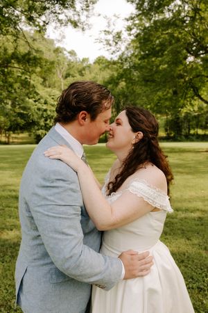 Bride and groom share a romantic almost-kiss moment outdoors, surrounded by green trees on their wedding day.