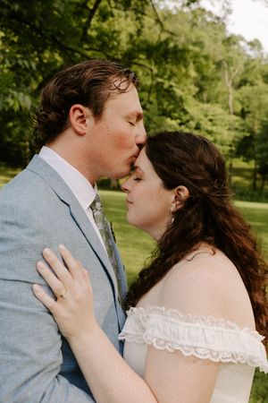 Groom in blue suit kisses bride on forehead as she closes her eyes in a serene outdoor setting.