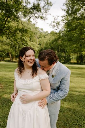 A laughing bride is embraced and kissed by her groom in a light blue suit during outdoor wedding portraits.