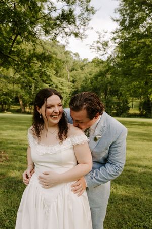 Groom in light blue suit whispering to laughing bride in white off-shoulder wedding gown on green lawn.