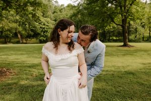 Bride in off-shoulder white gown smiles as groom in light blue suit whispers in her ear outdoors.